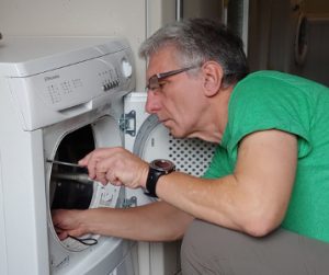 Apie meistrą, meistras, vilkintis žalią palaidinę, priklaupęs atsuktuvu suka varžtą skalbimo mašinos durelių zonoje
